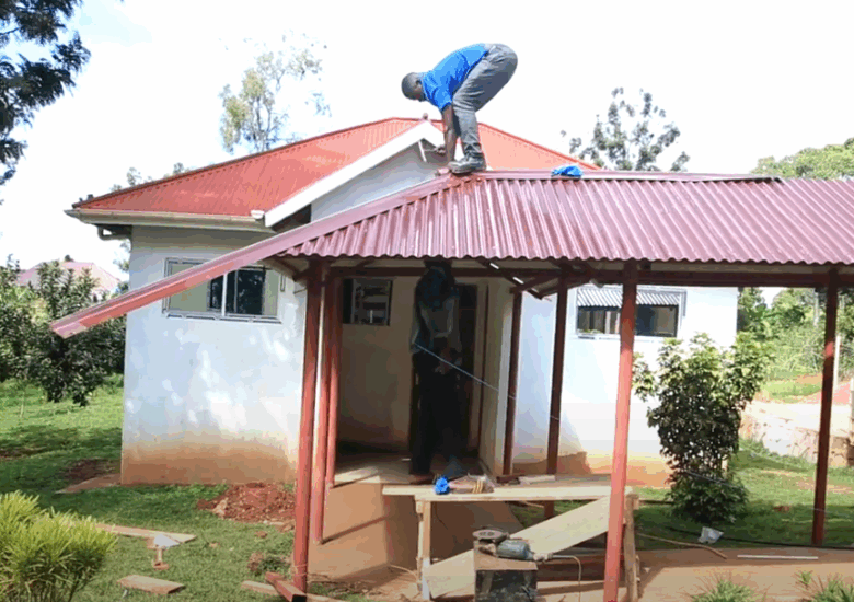 A person stands on the tin roof of a sheltered walkway, hunched over working on the roof, while another person stands below